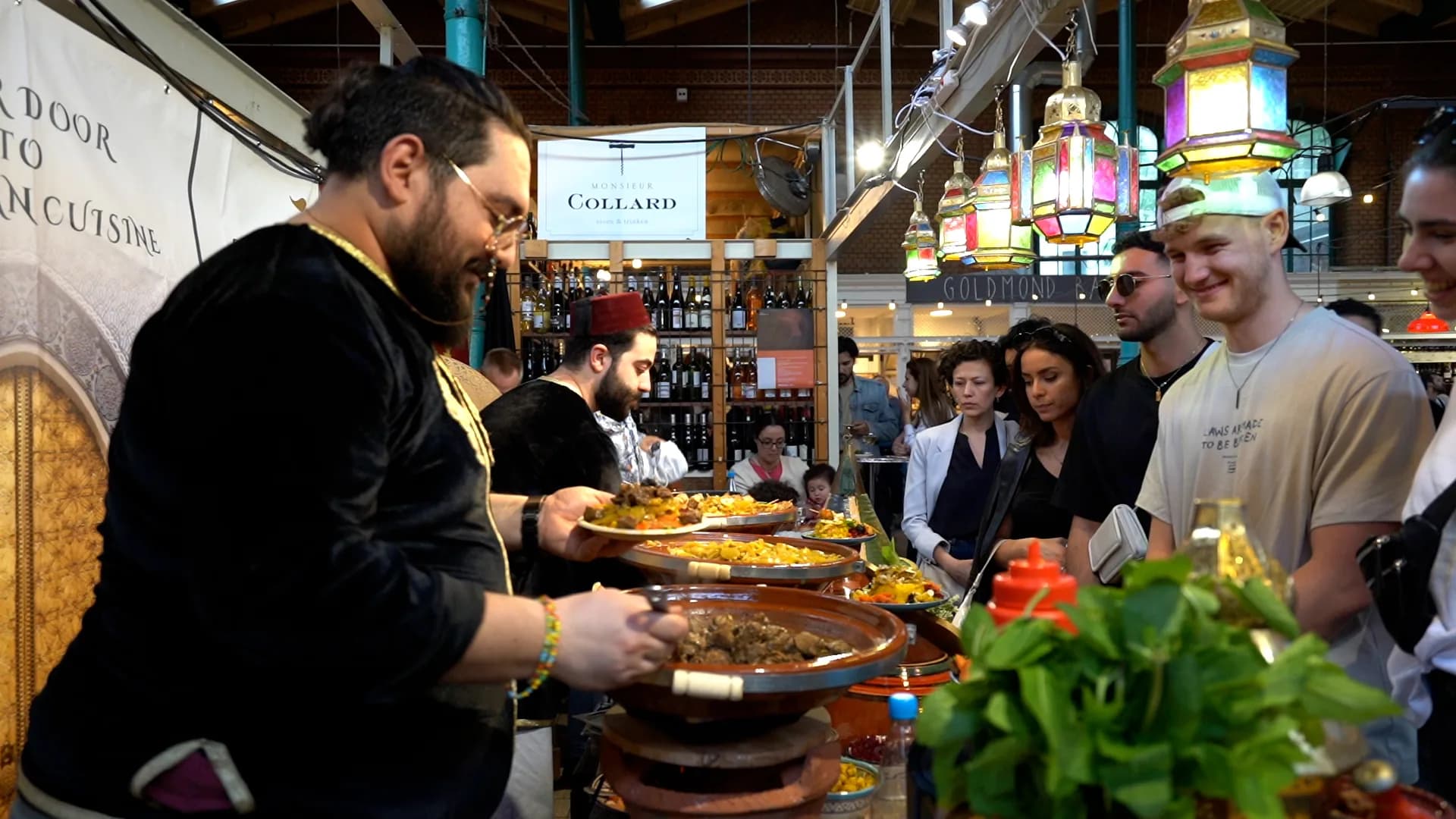 Ein Koch schöpft aus einer Tajine, während Gäste am Stand warten; darüber hängen marokkanische Laternen